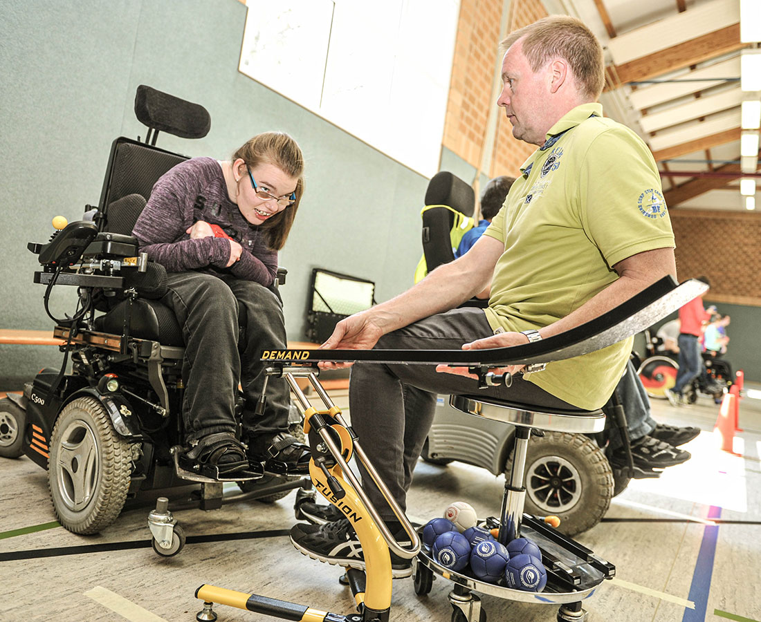 Betreuer hilft Frau im Rollstuhl bei Übung mit Bällen in Sporthalle